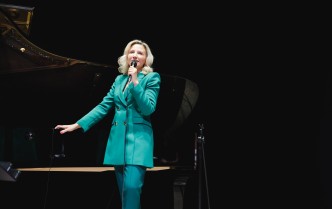 kobieta w zielonym garniturze trzyma mikrofon lewą ręką śpiewając, prawą ręką opierając się o kant fortepianu