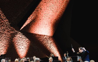 grupa muzyków w dolnej części zdjęcia ukazana wraz z dyrygentem, w górnej części oświetlona architektura sali koncertowej