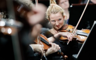 zbliżenie na kobietę grającą na skrzypcach, otoczona przez rozmyte postacie innych muzyków