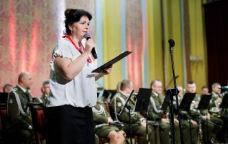kobieta w białej bluzce i czerwonych koralach na tle orkiestry wojskowej