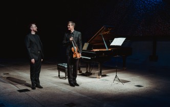 dwoch strojących mężczyzn obok fortepianu, jeden z nich trzyma skrzypce