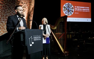 mężczyzna ubrany w czarny garnitur przemawiający ze sceny, obok niego kobieta ubrana w czerń i biel