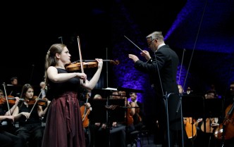 kobieta grająca na skrzypcach obok stoi dyrygen z podniesionymi rękami, w jednej ręce trzyma batutę