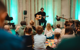 mężczyzna grający na gtarze, przed nim siedzą dzieci