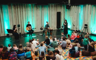 artyści na scenie oraz widownia - dorośli i dzieci siedzący na kolorowym dywanie