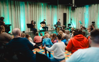 artyści na scenie oraz widownia - dorośli i dzieci siedzący na kolorowym dywanie
