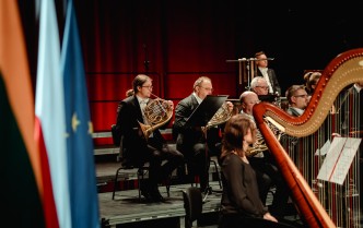 kobieta siedzi przy harfie, a nią siedzą muzycy grający na waltorniach