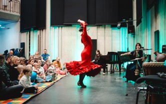 tańcząca kobieta w czerwonej sukni, po prawej stronie kobieta grająca na skrzypcach, po lewej rodzice i dzieci siedzący na kolorowym dywanie