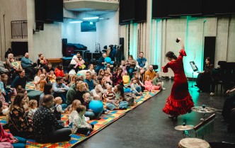 tańcząca kobieta w cerwonej sukni, oraz dorośli i dzieci siedzący na kolorowym dywanie
