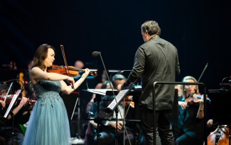 kobieta grająca na skrzypcach, po prawej zwrócony plecami stoi dyrygent