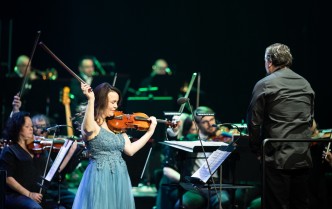 kobieta grająca na skrzypcach, po prawej zwrócony plecami stoi dyrygent