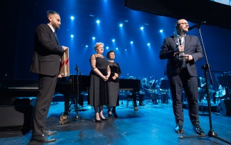 dwóch stojących mężczyzn w garniturach, pomiędzy nimi stoi kobieta w czarnej sukni