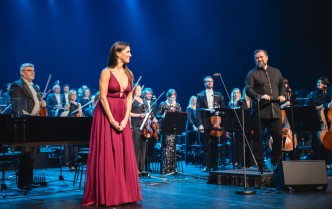 kobieta w czerwonej sukni, po prawej stronie stoi dyrygent, za nimi stoi orkiestra