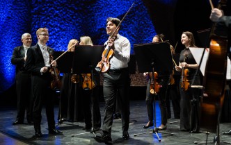 mężczyzna w białej koszuli trzyma skrzypce, w tle grupa osób w czarnych strojach trzymająca skrzypce