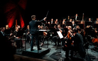 orkiestra siedzi na scenie, przed nią stoi dyrygent z uniesioną w ręce batutą