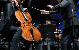mężczyzna w czarnym garniturze gra na wiolonczeli, po prawej stronie stoi mężczyzna w czarnym garniturze, który trzyma w ręce batutę