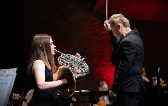 kobieta gra na waltorni, obok stoi dyrygent z uniesioną batutą