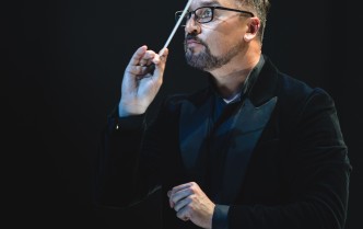 mężczyzna w okularach ma uniesioną rękę, w której trzyma batutę