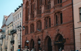 grupa zwiedzających osób stojąca przed budynkiem Dworu Artusa w Toruniu, całość ukazana z dalszej odległości tak, że widać wieżyczki budynku