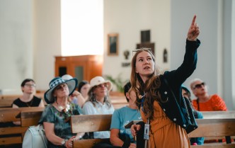 na pierwszym planie stojąca kobieta ukazana od pasa w górę z podniesioną dłonią w górę wskazująca na coś, za nią grupa zwiedzających siedząca w kościelnych ławkach