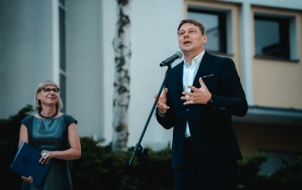 mężczyzna ubrany w czarny garnitur przemawia do mikrofonu, stoi w centralnej części fotografii, po lewej stronie stoi kobieta w szarej sukience, obie postacie ukazane od połowy pasa w górę