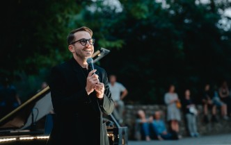 Mężczyzna w garniturze, solista, stoi i trzymając w dłoni mikrofon przemawia do publiczności, w tle za nim stoi fortepian