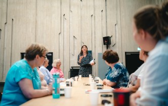 grupa kobiet siedząca przy stole, w centralnej części stół, na końcu którego stoi prowadząca warsztaty