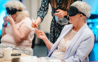 dwie kobiety siedzące przy stole, po lewej stronie wącha zapach, kobieta po lewej wypowiada się trzymając zapach w jednej dłoni, pomiędzy nimi stoi kobieta podająca zapachy