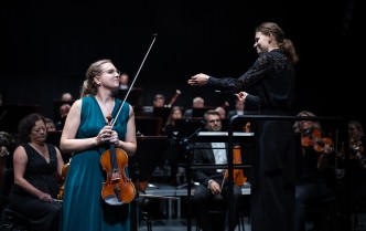 kobieta trzymająca skrzypce, obok kobieta z uniesinymi rękoma, w prawej dłoni trzyma batutę