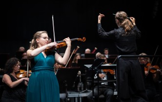 kobieta grająca na skrzypcach, obok kobieta z uniesinymi rękoma, w prawej dłoni trzyma batutę