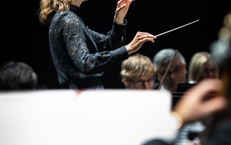 kobieta w uniesionymi rękoma, w jednej dłoni trzyma batutę