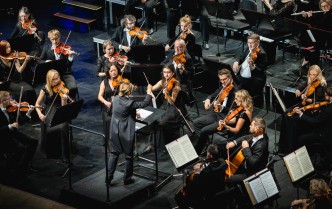 grupa osób grających na instrumentach smyczkowych, po środku stoi dyrygentka