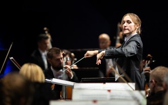 kobieta z uniesionymi rękoma, w prawej dłoni trzyma batutę