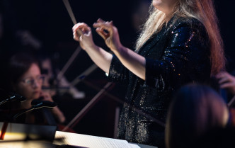 kobieta z uniesionymi rękoma, w jednej dłoni trzyma batutę - dyrygentka