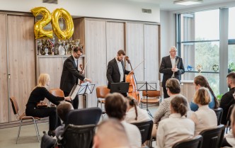 siedząca kobieta i trzecha stojących mężczyzn, trzumają instrumenty smyczkowe, przed nimi siedzi tyłem kilka osób