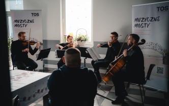 jedna osoba zwrócona tyłem siedzi przed czworgiem muzyków grających na instrumentach smyczkowych