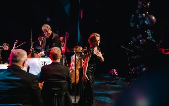 uśmiechnieta kobieta, po lewej stronie siedzą mężczyźni grajacy na skrzypcach