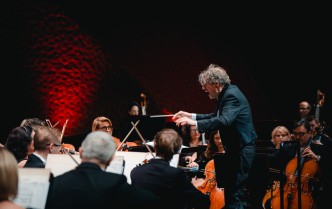 mężczyzna trzymający ręce w górze, w prawej dłoni trzyma batutę