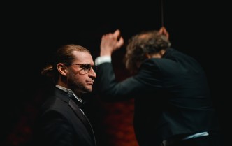 mężczyzna w garniturze, za nim tyłem mężczyzna z uniesionymi do głowy rękoma