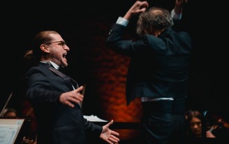 mężczyzna w garniturze z rozłożonymi rękoma, za nim tyłem mężczyzna z uniesionymi do głowy rękoma