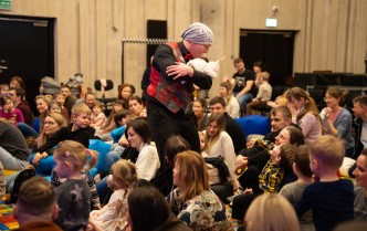 dzieci i dorośli siedzą na dywanie, wśród nich stoi mężczyzna przebrany za krasnala