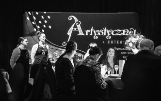 kobiety i mężczyźni siedzący przy stole, z boku stąją kelnerki, w tle napis Artystyczna