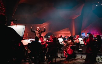 siedzący tyłem muzycy grający na instrumentach, przodem stoi dyrygent z uniesionymi rękoma