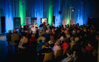 publiczność na widowni, przed nimi siedzą przodem kobieta przy fortepianie, oraz kobieta i mężczyzna