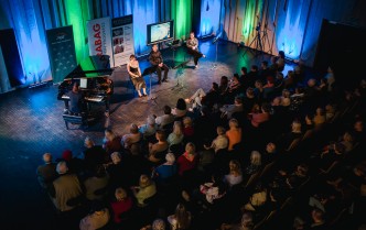 publiczność na widowni, przed nimi siedzą przodem kobieta przy fortepianie, oraz dwie kobiety i mężczyzna siedzą na krzesłach