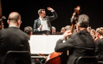 mężczyzna stojący przodem z uniesionymi rękoma, w prawej dłoni trzyma batutę, przed nim siedzą muzycy