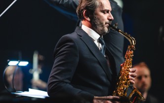 mężczyzna grający na saksofonie tenorowym, za nim stoi dyrygent z uniesioną ręką, w dłoni trzyma batutę