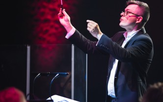 mężczyzna z uniesiony w górę rękoma, w prawej ręce trzyma batutę