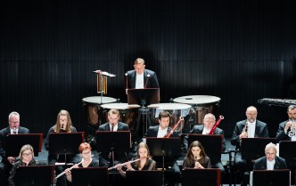grupa osób grających na instrumentach dętych drewnianych, za nimi mężczyzna grający na kotłach