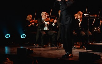 mężczyzna śpiewający do mikrofonu, w tle siedzi orkiestra